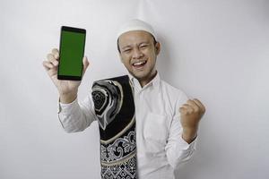 A happy young Asian Muslim man with a successful expression showing copy space on his phone isolated by white background photo