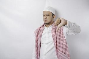 Disappointed Asian Muslim man gives thumbs down hand gesture of approval, isolated by white background photo