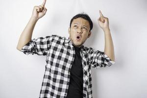 Shocked Asian man wearing tartan shirt pointing at the copy space on top of him, isolated by a white background photo