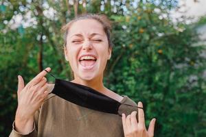 Happy positive girl takes off protective medical mask from face outdoors. Young woman removing mask smiling after vaccination. Coronavirus pandemic Covid 19 concept. Spring flowers pollen allergy. photo