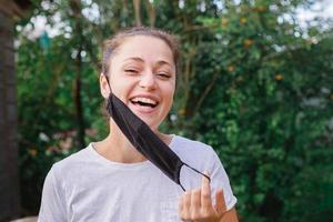 Happy positive girl takes off protective medical mask from face outdoors. Young woman removing mask smiling after vaccination. Coronavirus pandemic Covid 19 concept. Spring flowers pollen allergy. photo