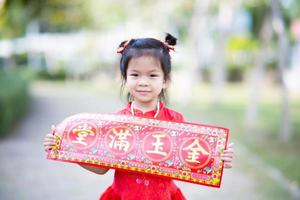 retrato linda chica asiática con tarjeta de felicitación de año nuevo chino que significa rico y feliz. los niños sonríen dulcemente. niño use cheongsam rojo en el jardín. niño de 5 años. foto