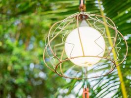 An old iron lantern with an orange glow illuminates a circle. It is located in a park with green leaves. photo