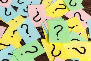 Pile of colorful paper notes with question marks on wooden background. Closeup photo