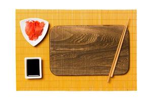 Empty rectangular brown wooden plate with chopsticks for sushi, ginger and soy sauce on yellow bamboo mat background. Top view with copy space for you design photo