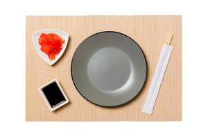 Empty round gray plate with chopsticks for sushi and soy sauce, ginger on brown sushi mat background. Top view with copy space for you design photo