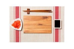 Empty rectangular brown wooden plate with chopsticks for sushi, ginger and soy sauce on sushi mat background. Top view with copy space for you design photo