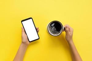 Female hands holding black mobile phone with blank white screen and mug of coffee. Mockup image with copy space. Top view on yellow background, flat lay photo