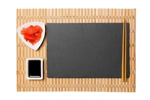 Empty rectangular black slate plate with chopsticks for sushi, ginger and soy sauce on yellow bamboo mat background. Top view with copy space for you design photo