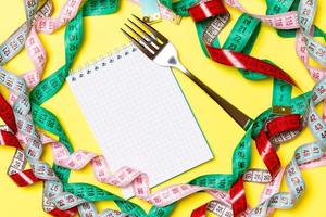 Top view of notebook with fork surrounded with colorful measuring tapes on yellow background. Copy space with diet planning photo