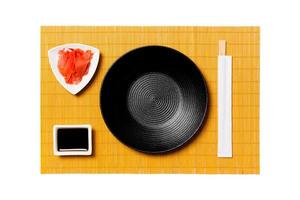 Empty round black plate with chopsticks for sushi and soy sauce, ginger on yellow bamboo mat background. Top view with copy space for you design photo