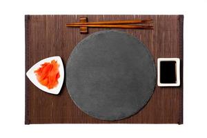 Emptyround black slate plate with chopsticks for sushi, ginger and soy sauce on dark bamboo mat background. Top view with copy space for you design photo