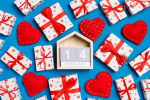 Composition of wooden calendar, holiday white gift boxes and red textile hearts on colorful background. The fourteenth of February. Valentine's day concept photo