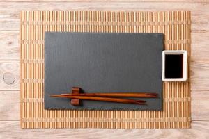 Empty rectangular black slate plate with chopsticks for sushi and soy sauce on wooden background. Top view with copy space photo