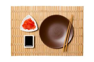 Empty round brown plate with chopsticks for sushi, ginger and soy sauce on yellow bamboo mat background. Top view with copy space for you design photo