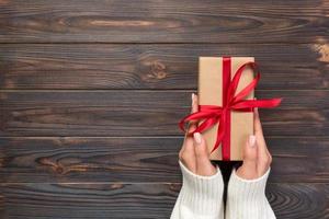 las manos de las mujeres dan San Valentín envuelto u otro regalo hecho a mano en papel con cinta roja. caja de regalo, decoración de regalo en mesa de madera negra, vista superior con espacio de copia foto