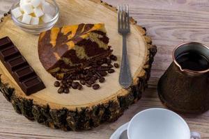 A coffee cup with a piece of cake on wooden stump, a chocolate bar, fork, , coffee beans and bowl with sugar cubes on a wooden table photo