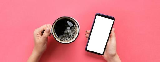 Female hands holding black mobile phone with blank white screen and mug of coffee. Mockup image with copy space for you design. Top view banner on pink background, flat lay photo