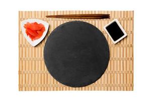 Emptyround black slate plate with chopsticks for sushi and soy sauce, ginger on yellow bamboo mat background. Top view with copy space for you design photo