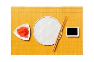 Empty round white plate with chopsticks for sushi and soy sauce, ginger on yellow bamboo mat background. Top view with copy space for you design photo