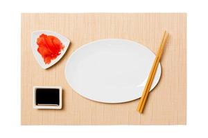 Empty oval white plate with chopsticks for sushi and soy sauce, ginger on brown sushi mat background. Top view with copy space for you design photo