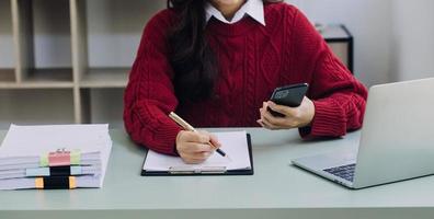 Young business woman sitting in office at table and using smartphone. On desk is laptop and tablet computer, on screen charts and graphs. Woman analyzing data. Student learning online. photo