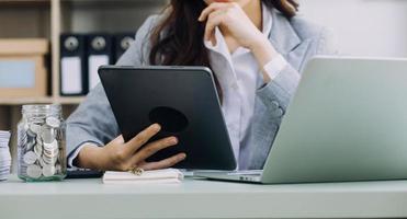 Young business woman sitting in office at table and using smartphone. On desk is laptop and tablet computer, on screen charts and graphs. Woman analyzing data. Student learning online. photo