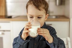 Little boy sitting in the kitchen and drinking milk. Fresh milk in glass, dairy healthy drink. Healthcare, source of calcium, lactose. Cozy and modern interior. Preschool child with casual clothing. photo