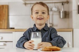 Little boy sitting in the kitchen and drinking milk. Fresh milk in glass, dairy healthy drink. Healthcare, source of calcium, lactose. Cozy and modern interior. Preschool child with casual clothing. photo