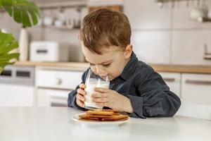 Little boy sitting in the kitchen and drinking milk. Fresh milk in glass, dairy healthy drink. Healthcare, source of calcium, lactose. Cozy and modern interior. Preschool child with casual clothing. photo