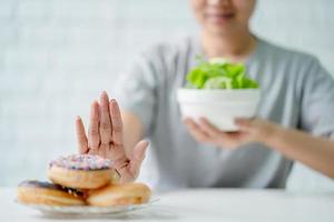 joven mujer o niña asiática empujando comida chatarra y donas dulces con las manos. ella elige vegetales de ensalada para una buena salud. foto