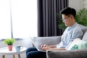 Side view of young Asian working near a large window and sitting on the sofa at home, With copy space. photo