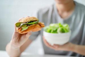 Closeup shot of fried chicken burger in hand, young woman on diet picking her healthy salad. Dieting and good health concept. photo