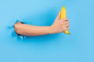 Hand through paper holding banana on blue background photo