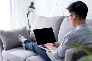 los asiáticos usan anteojos, trabajan en una computadora portátil mientras están acostados en el sofá en casa. foto
