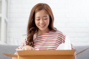 una mujer asiática feliz y sonriente está sacando cosas de una caja ordenada por Internet. concepto de compras en línea foto