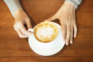 people hand holding hot cup of coffee with flower tree shape foam over ready to drink on rustic wooden table, top view. filter tone vintage style. photo