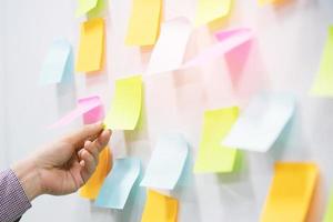 primer plano mano personas hombre de negocios bloc de notas en la pared en la sala de reuniones. tablero de programación de recordatorio de papel de nota de publicación adhesiva. espacio vacío de copia de variedad colorida. enfoque suave. foto