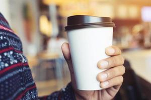 mano de hombre joven sosteniendo una taza de papel para llevar bebiendo café caliente en la cafetería. foto