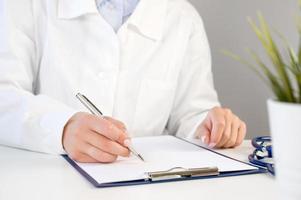 primer médico en una bata blanca escribiendo una receta o llenando el formulario en el gabinete del hospital. concepto de atención médica foto