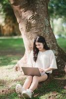 Asian woman sitting on picnic and lawn at park working on laptop. Asian female using laptop while sitting under a tree at park with bright sunlight. Work from anywhere concept photo