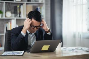 Asian Business man wear glasses and hand over head having stressful depression sad time while working on laptop at home. Depression man sad serios working from home. Working at anywhere concept. photo