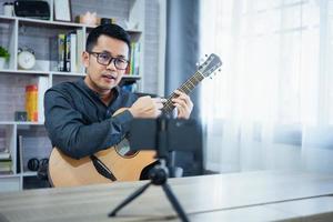 Asian music teacher man broadcasting live guitar lessons to students online using mobile. Asian male musician playing guitar singing live video conferencing at home. Live concert music from anywhere. photo