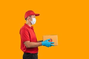 Side view. Asian delivery man worker wearing surgical mask and medical gloves in red uniform isolated on yellow background, hold parcel boxes for sending or conveying parcels by mail. photo