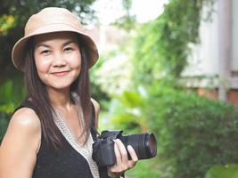 mujer asiática, con sombrero y blusa negra sin mangas, parada en el jardín y sosteniendo una cámara dslr, sonriendo alegremente. foto