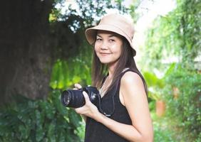mujer asiática, con sombrero y blusa negra sin mangas, parada en el jardín y sosteniendo una cámara dslr, sonriendo alegremente. foto
