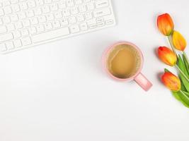 flat lay of computer keyboard, pink cup of coffee and tulips on white background with copy space. photo