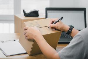 cierre la mano un hombre use un bolígrafo escriba en la caja del paquete sobre la dirección de entrega del paquete y use una computadora portátil para registrarse en el sistema en línea foto
