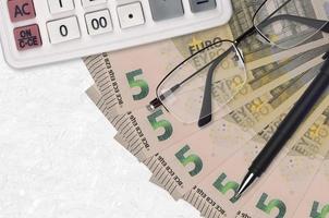 Ventilador de billetes de 5 euros y calculadora con gafas y bolígrafo. préstamo comercial o concepto de temporada de pago de impuestos foto