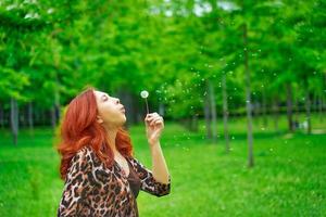 Cute red-haired woman in leopard jacket blows away a dandelion. photo